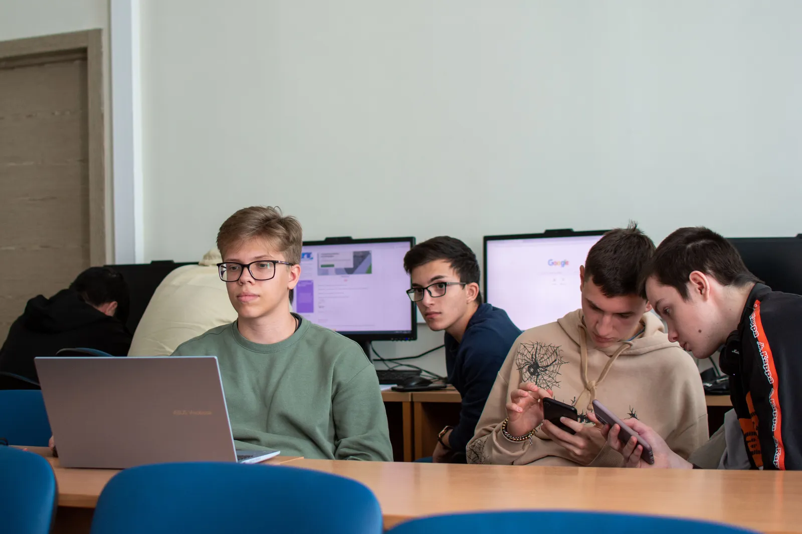 Four students at classroom computers working on a project, with one using a laptop in the foreground.